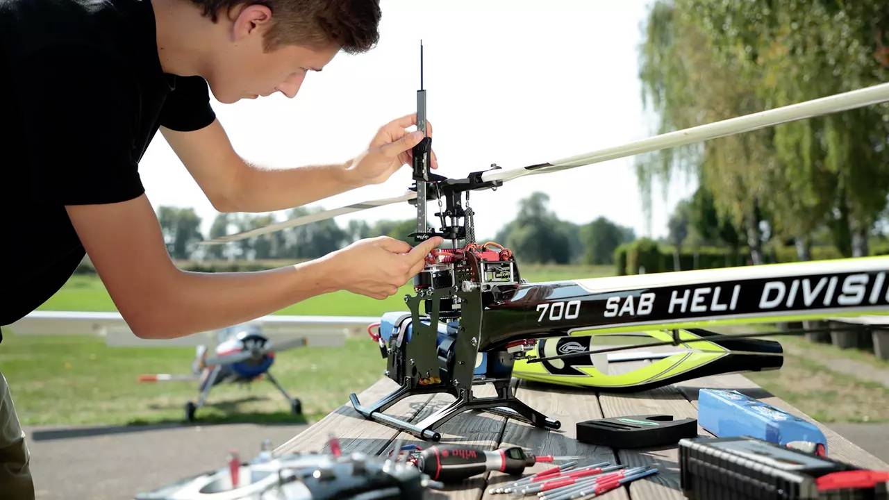 A man is working outdoors on a model helicopter with the inscription 'SAB Heli Division'. Tools and accessories are laid out on the table.