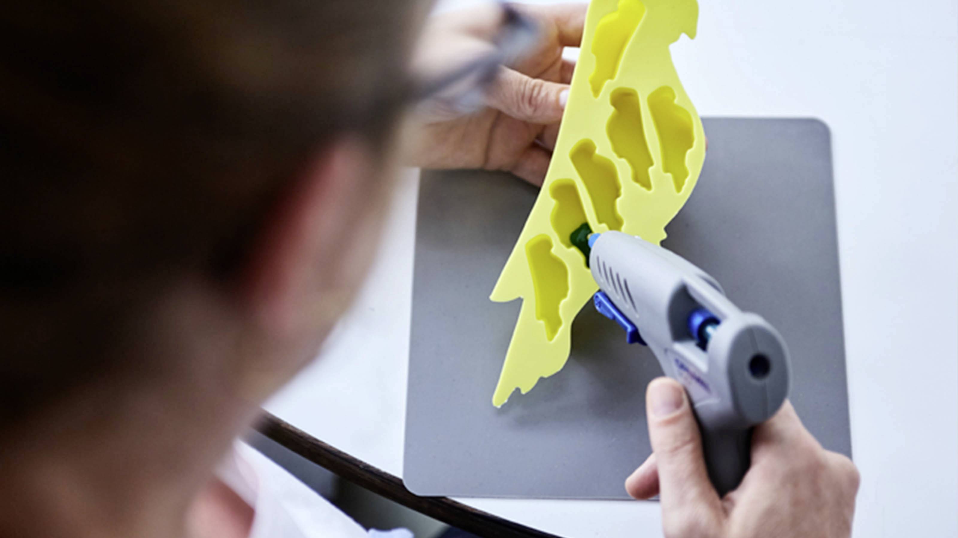 A person is forming a yellow foam material using a hot glue gun on a grey surface.