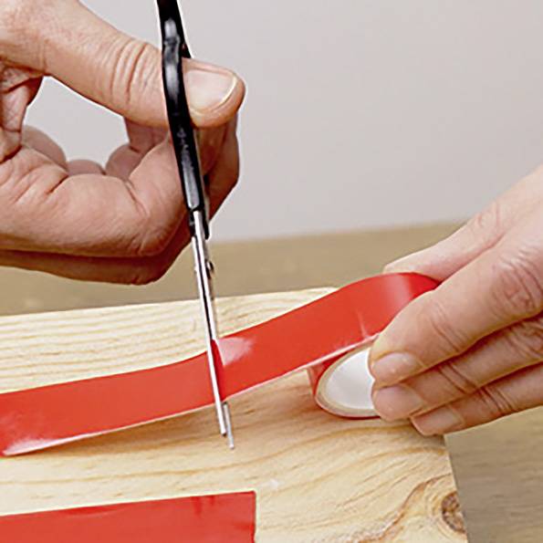 A person is cutting a strip of red adhesive tape on a wooden surface using scissors.