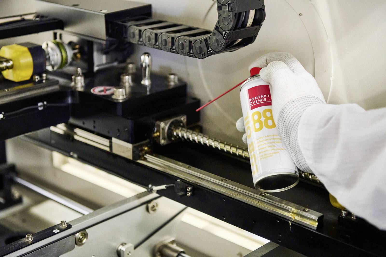 A person in protective clothing is spraying lubricant from a red can onto an industrial machine with metal components, presumably for maintenance purposes.