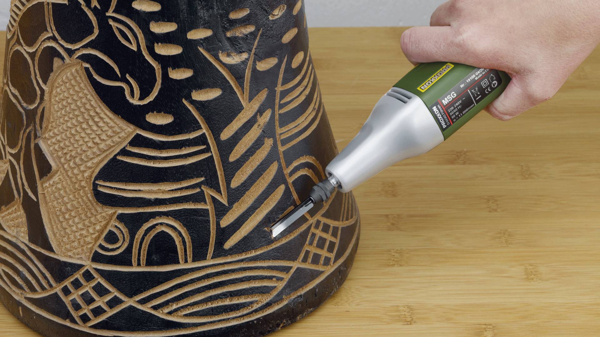 A hand operates an engraving tool on a wooden object with tribal patterns, illustrating precise craftsmanship on a wood surface.