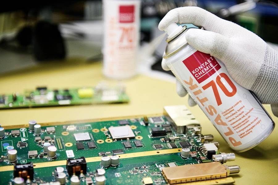 A gloved hand sprays 'Plastik 70' onto a green circuit board, suggesting protection or maintenance of the electronic components.