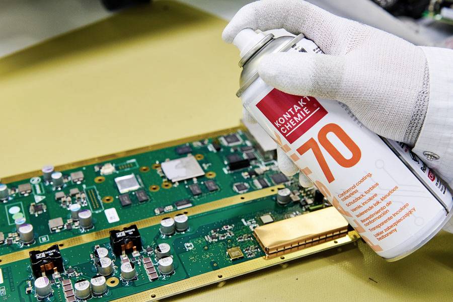 A gloved hand sprays electronic cleaner onto a green circuit board on a workbench, showing maintenance of electronic components.