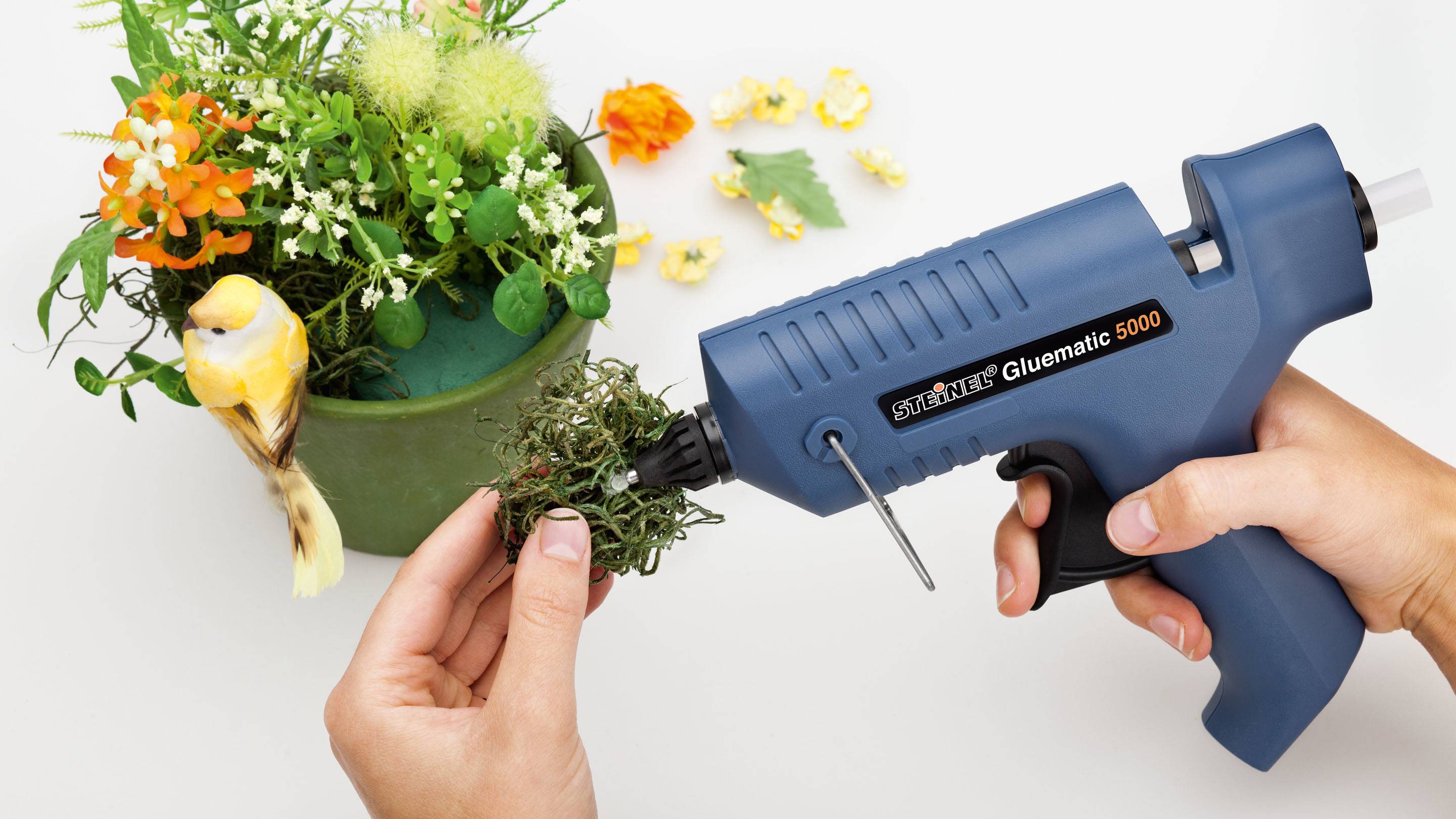 A person is using a blue hot glue gun to secure artificial plants and decorative elements in a green planting pot.