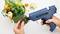 A person is using a blue hot glue gun to secure artificial plants and decorative elements in a green planting pot.