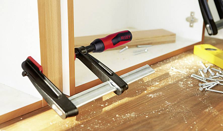 A G-clamp holds two wooden boards together. On a workbench, there are wood shavings, screws, and a yellow tool.