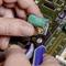 A person is placing a green battery onto an electronic circuit board using pliers and connecting it with a wire.