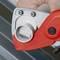 Close-up of a red pipe cutter in action, cutting a metal pipe. A hand is holding the tool.