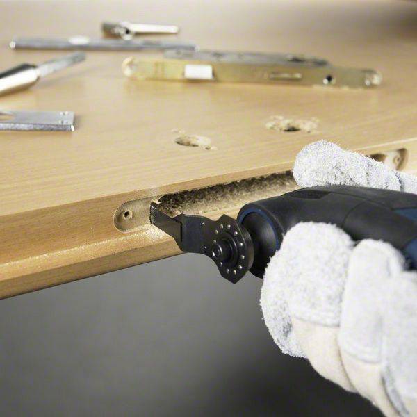 A person wearing a glove is operating an electric drill to bore a hole in a wooden door. Tools are lying beside the door.