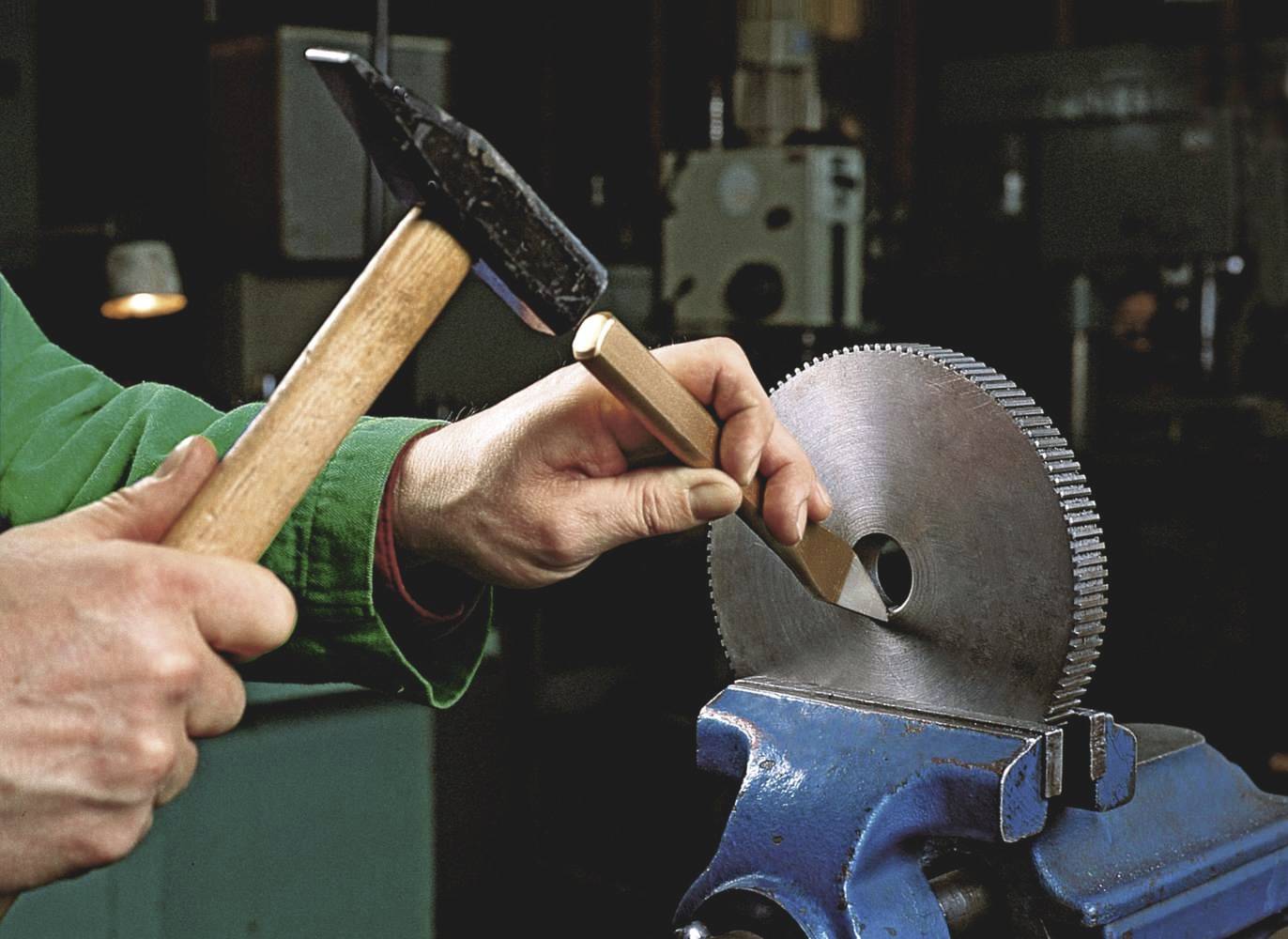 A person is working on a metallic gear with a hammer and chisel in a workshop. The focus is on precise manual craftsmanship.