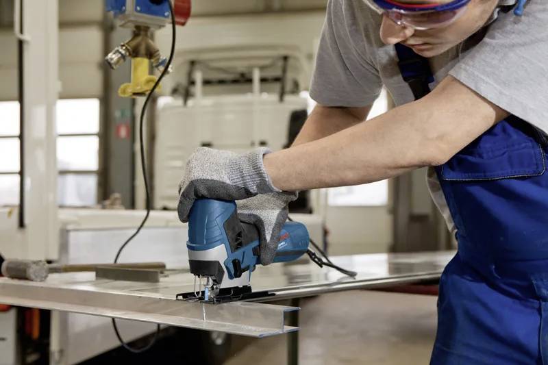 A person in safety gear uses an electric jigsaw to cut a sheet of metal in a workshop, concentrating on precise cutting.