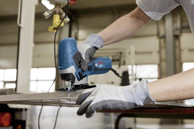 A person uses a blue jigsaw tool to cut a piece of metal in a workshop, wearing gray gloves for safety.