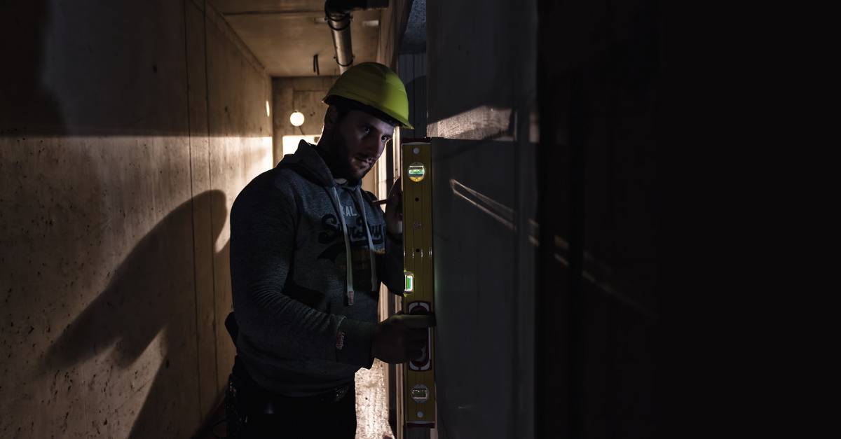 A construction worker wearing a yellow hard hat is using a spirit level to check a wall in a poorly lit tunnel.
