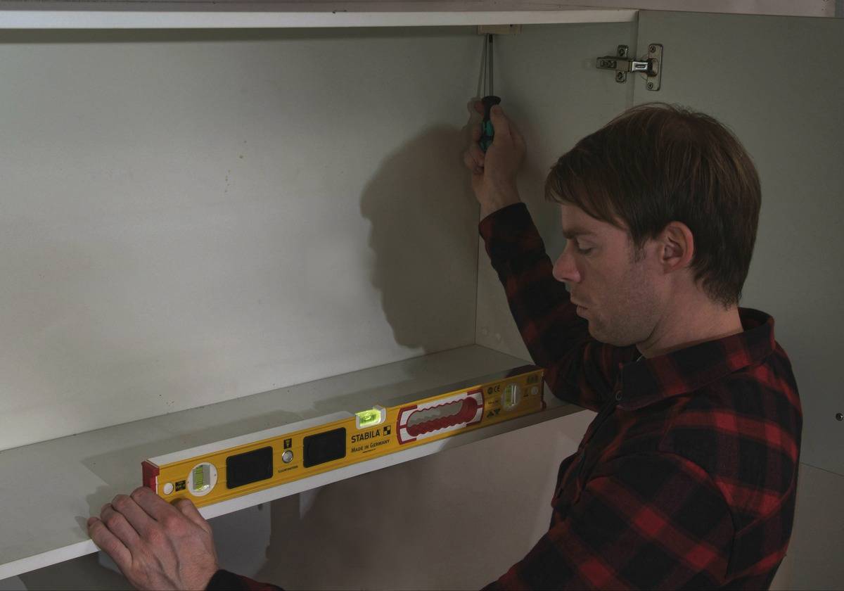 A man is installing a shelf, using a spirit level and a screwdriver.
