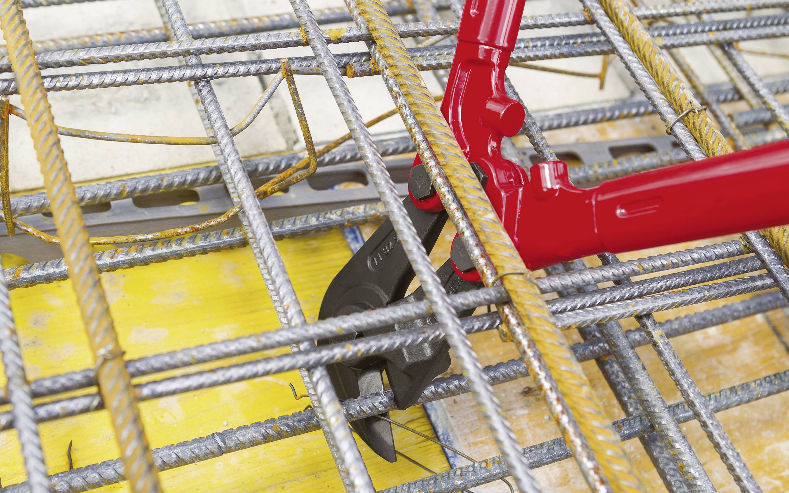 A red bolt cutter slices through thick metal struts in a construction site setting.