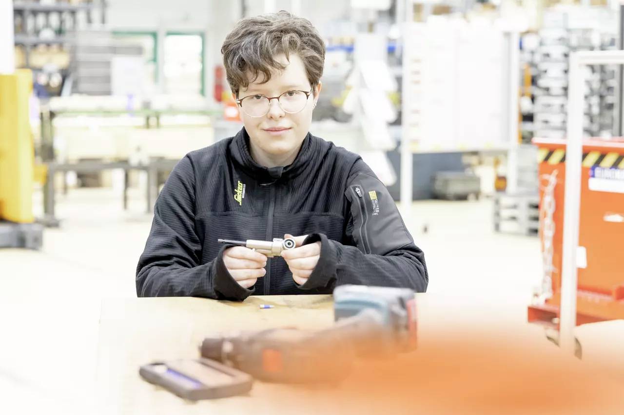 A person wearing glasses and a black jacket is seated at a table, holding a small tool, in a brightly lit workshop setting.