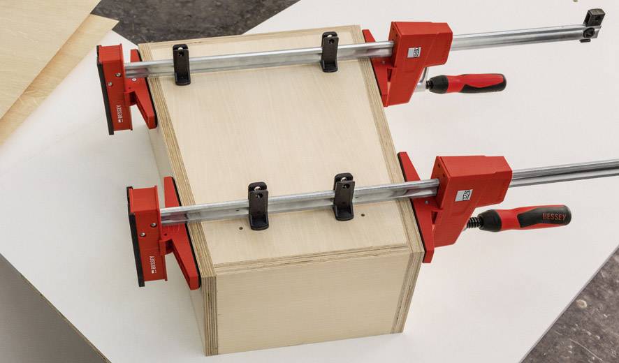 Two red G-clamps are holding a wooden box together. They are attached to a workbench and securing the assembled wood.