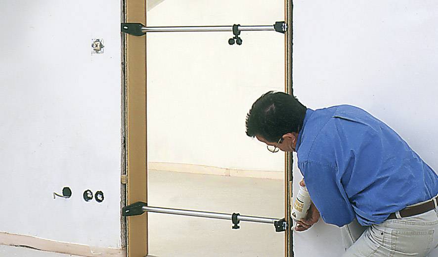 A man in a blue shirt and glasses is securing a door to a frame using support struts. A mitre saw lies on the floor.