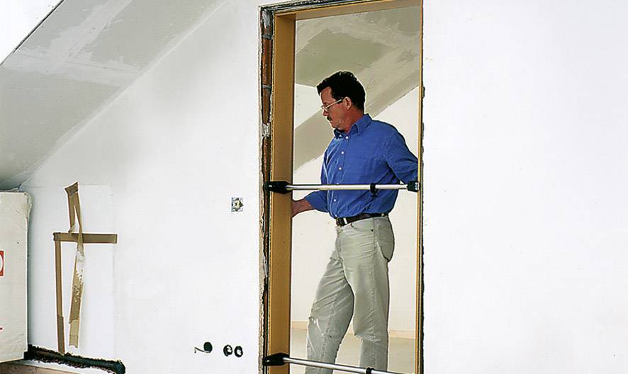A man in blue workwear is measuring a door opening in a white room using a measuring pole.