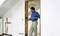 A man in blue workwear is measuring a door opening in a white room using a measuring pole.