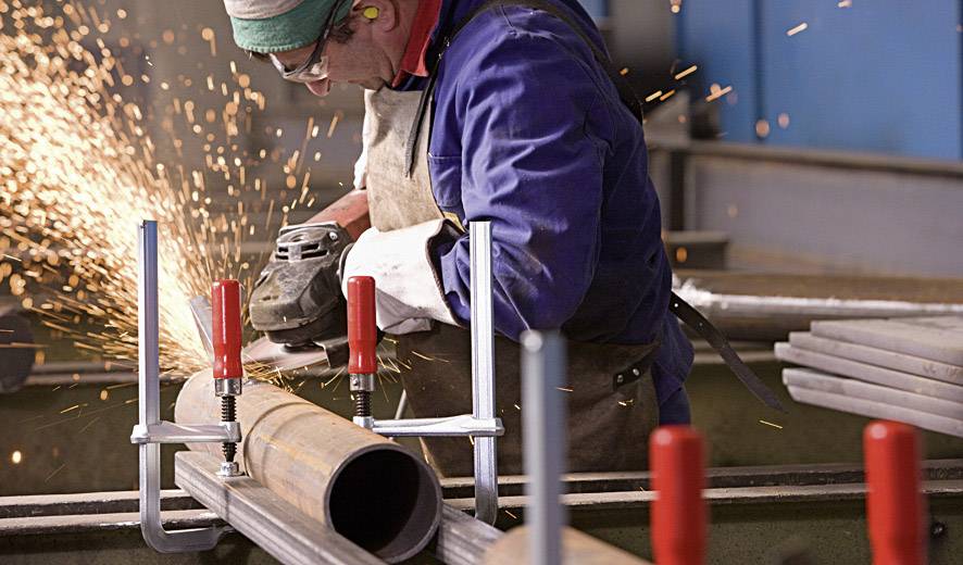 A person wearing protective equipment is grinding a metal pipe with an angle grinder in a workshop, sparks flying.