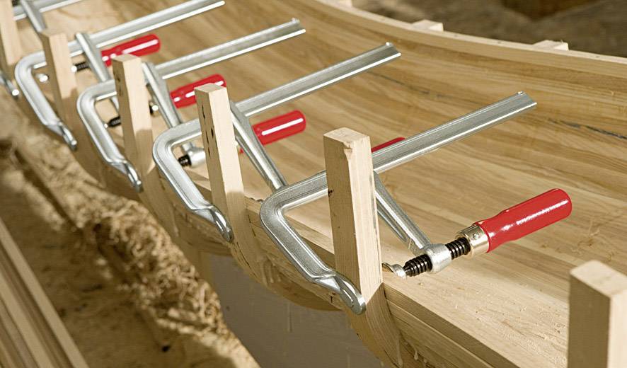 Several G-clamps hold curved wooden planks together, which are being used for construction.