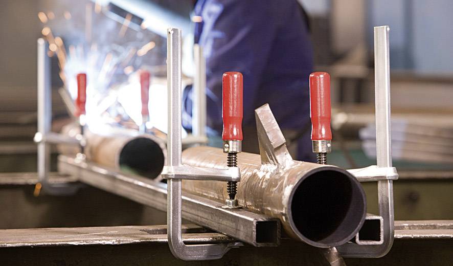 A person is welding two metal pipes together, held by red clamps, in a workshop environment. Sparks are flying from the welding.