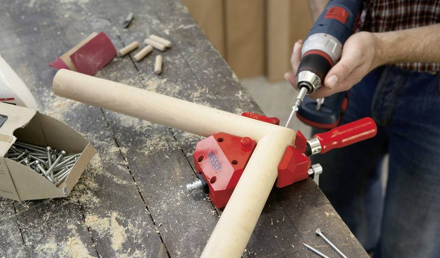 A person is using a drill to connect two wooden rods on a wooden table. Screws and tools are visible.