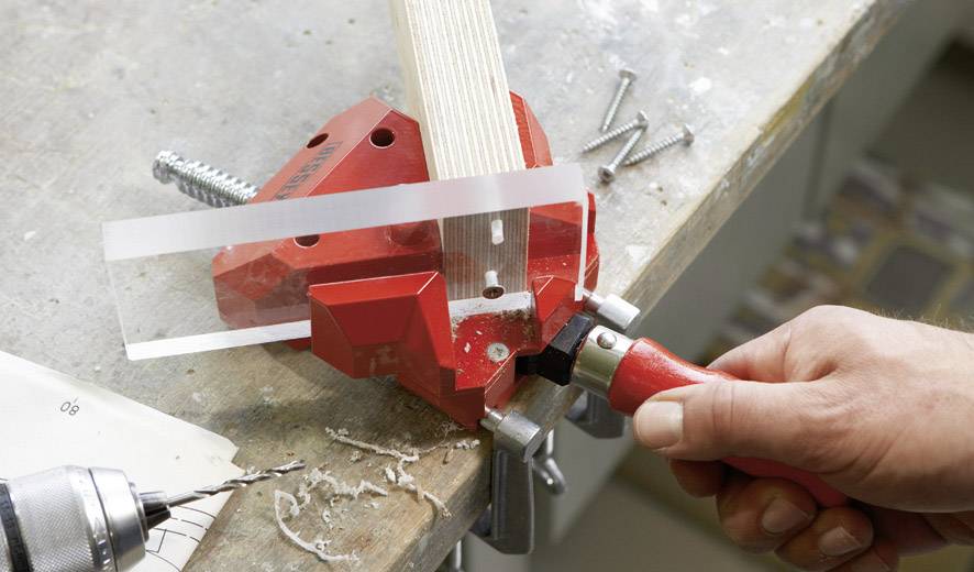A hand is using a red corner clamp tool to hold a piece of wood on a table. Screws and a drill are lying nearby.