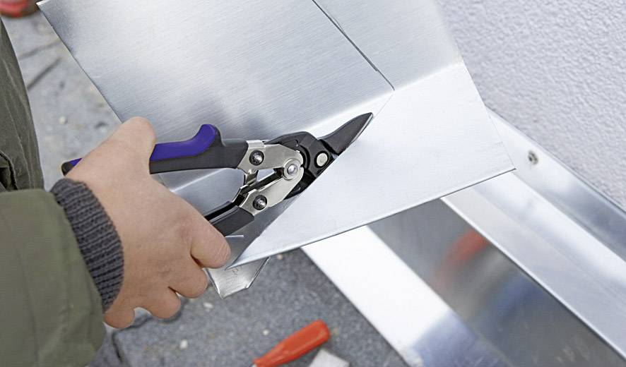 A person is cutting metal sheet with tin snips. The scene shows detailed work on a metal edge.