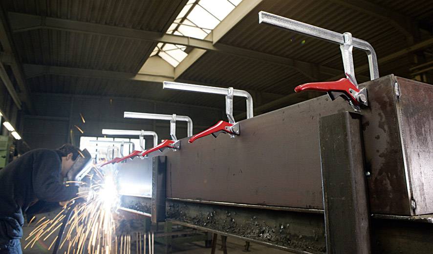 A metalworker is welding a large metal piece in a factory hall. Sparks are flying. Large red G-clamps are holding the workpiece firmly in place.
