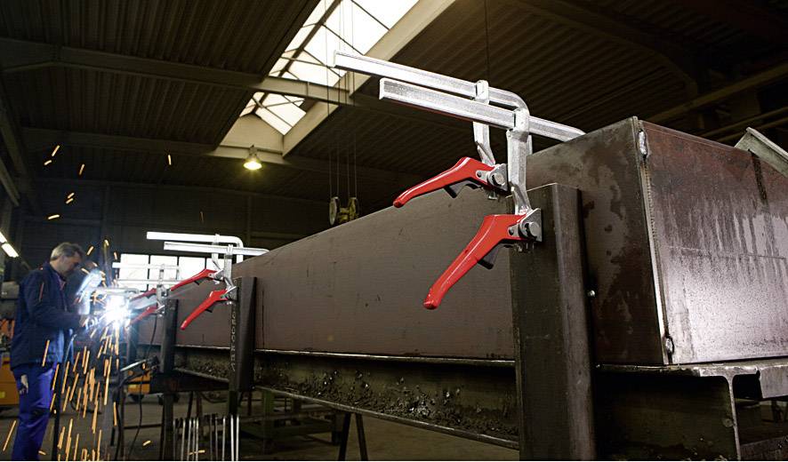 A person is welding in a workshop, sparks flying through the air. Large metal clamps are attached to a workpiece.