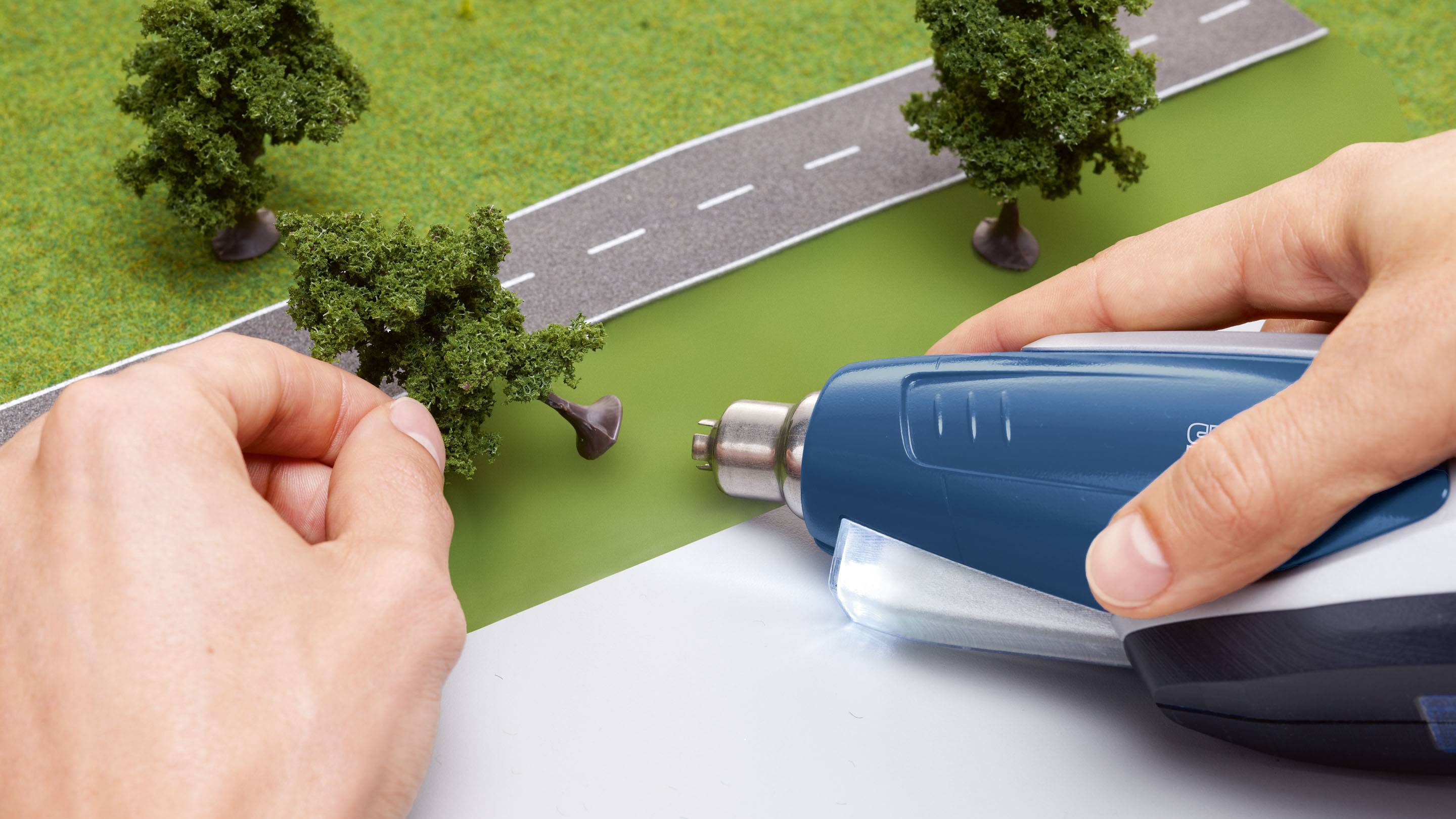 A hand holds a tree, another hand uses a tool to secure it on a road model.
