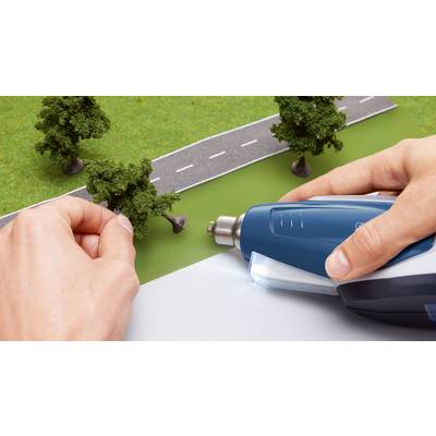 A hand holds a tree, another hand uses a tool to secure it on a road model.