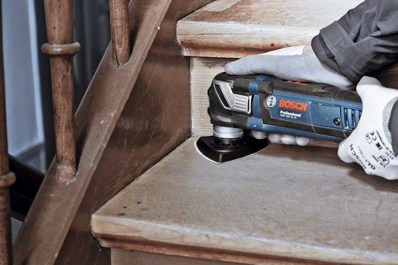 A person is using an oscillating power tool to sand a wooden staircase. The device is made by Bosch.