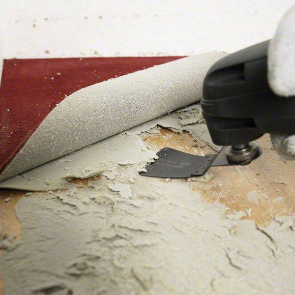 An electric grinding tool removes old carpet adhesive from a wooden floor, with a corner of the carpet folded up.