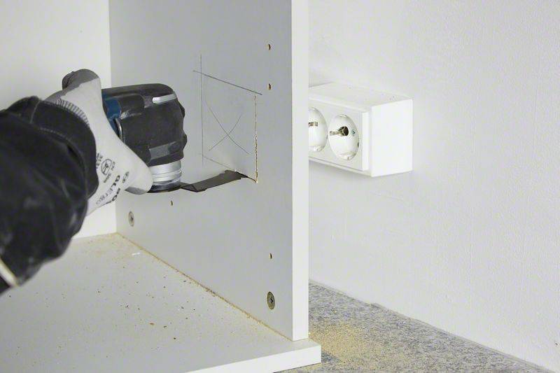 A person is cutting a square hole in the back of a white cabinet using an electric saw. A power socket is visible on the right.