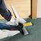 A person is using an electric tool to cut a grey section of pipe in front of a wooden door on a green carpet.