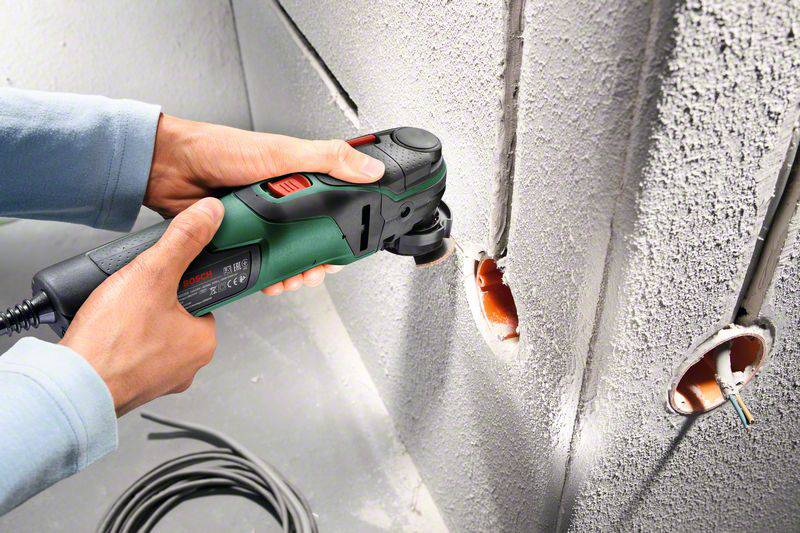 A person is using an electric multi-functional tool to drill holes in a concrete wall. Cables are visible.