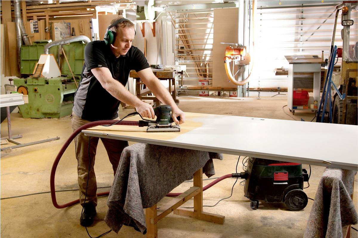A man is grinding a large wooden board in a workshop. He is wearing ear protection and is focused on his work.