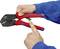 Two hands are holding a red and black crimping tool. One hand is pointing to a lever on the pliers. The person is wearing a blue jumper.
