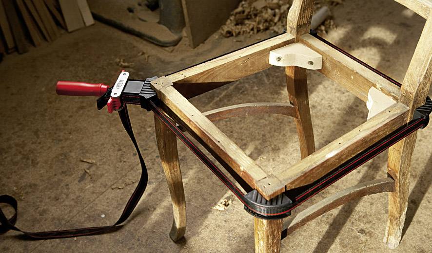 Wooden chair in the repair process, held in place with a red and black clamp to secure the legs. Wood shavings in the background suggest a workshop setting.