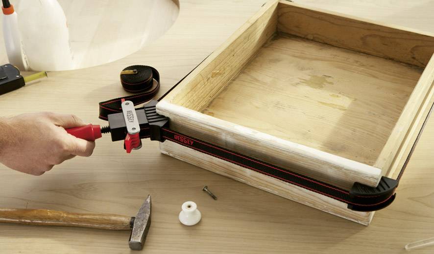 A hand pulls a box tight with a tension band frame. Beside it, a hammer, a screw, and wooden dowels lie on a table.
