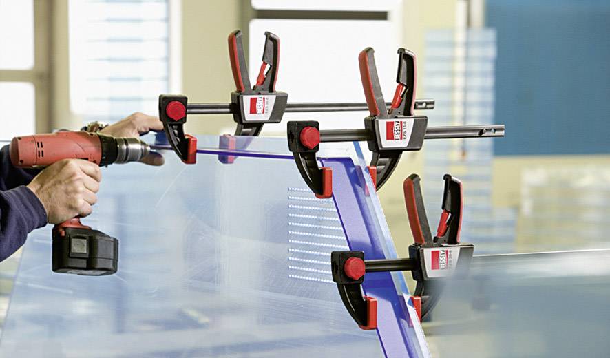 A person is working on a glass pane with an electric drill, which is stabilised by red G-clamps.