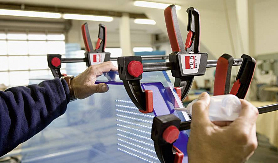 A person is using red clamps to hold glass panels together in a workshop. Hands are adjusting and securing the panels.