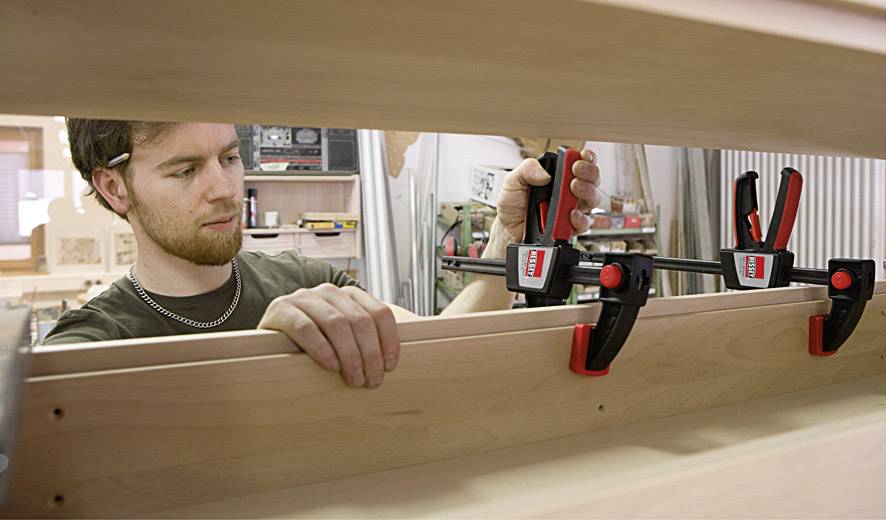 A man is working in a workshop and securing wooden planks with large clamps. Tools and woodwork can be seen in the background.
