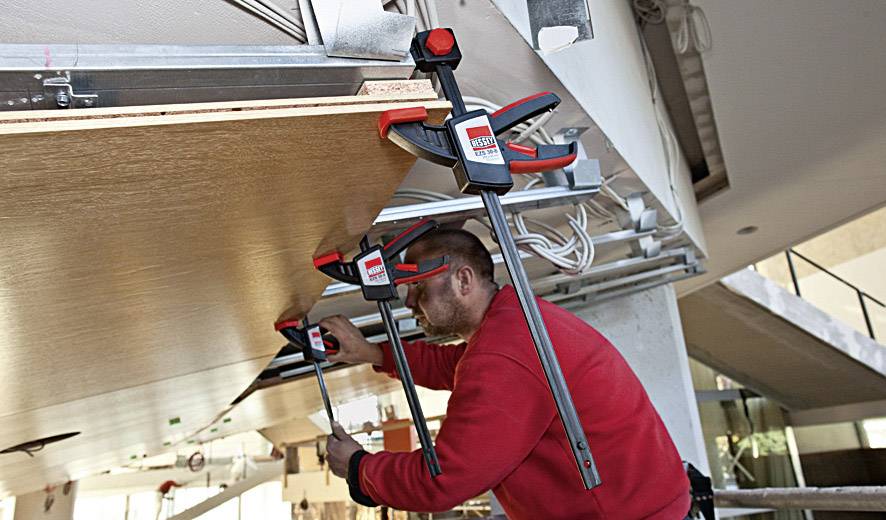 A craftsman is securing a wooden board to the ceiling with large clamps in a modern building.