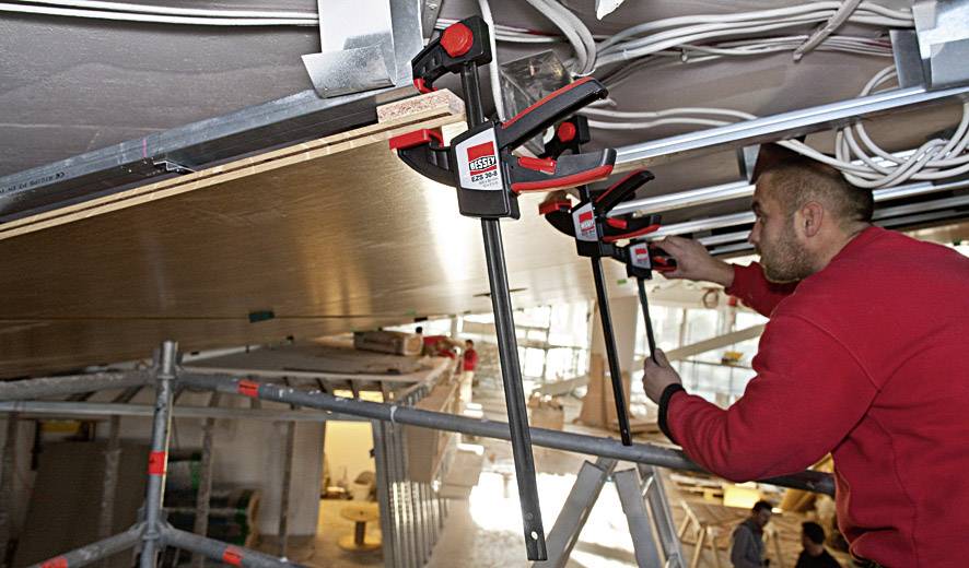 A man in red workwear is securing a plate to a ceiling using large clamps, surrounded by cables and scaffolding.