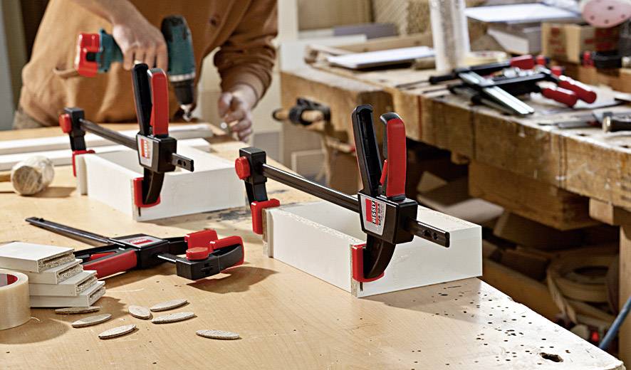 A person in a workshop secures wood with black and red G-clamps on a workbench, preparing another workpiece.
