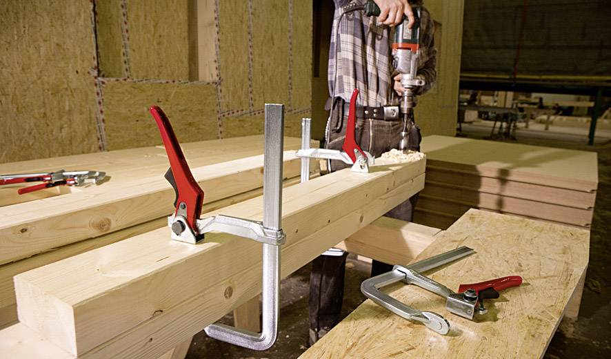 A person is drilling a wooden beam that is secured with two red clamps. Wooden boards are visible in the background.